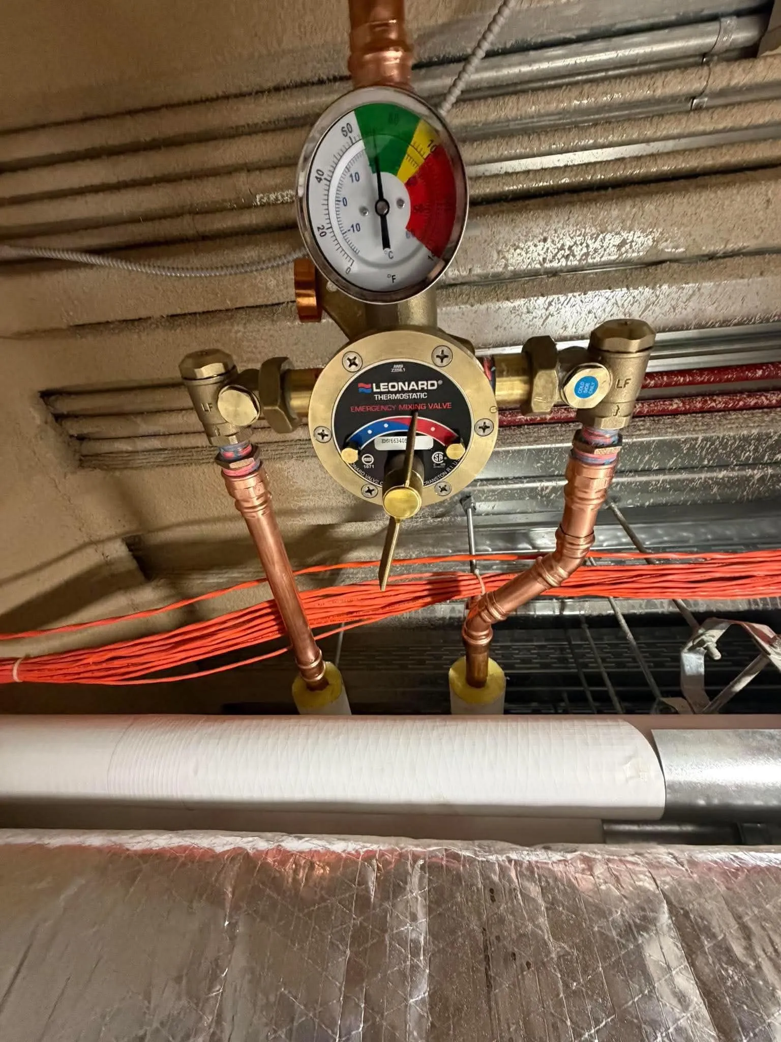 Gros plan d’un mitigeur thermostatique d’urgence : thermomètre en zone verte, raccords de cuivre sertis et vannes d’arrêt de chaque côté